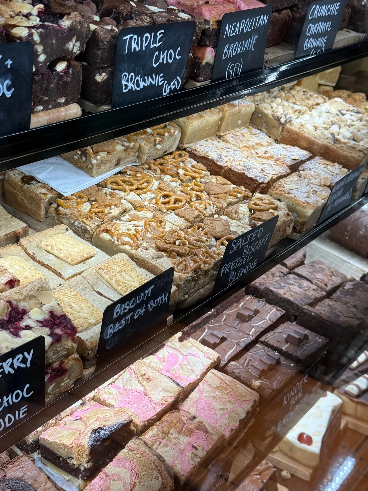 Um balcão de vidro com sobremesas em Bath empilhadas com brownies e blondies, incluindo brownie de triplo chocolate e blondie de pretzel Biscoff salgado, com rótulos em quadro-negro.