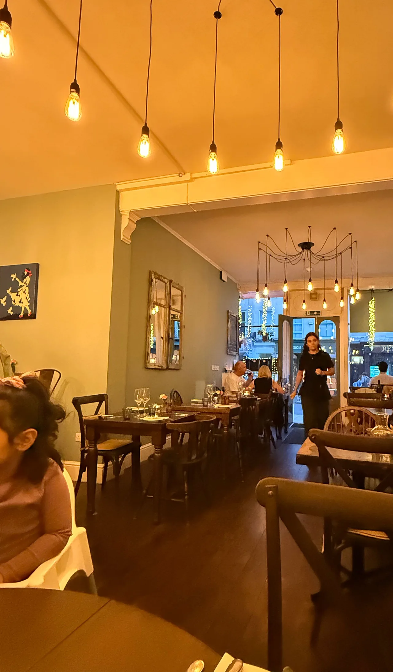 A sala de jantar da Chez Dominique em Bath, com paredes verde-sálvia, mesas de madeira escura e lâmpadas nuas penduradas do teto.