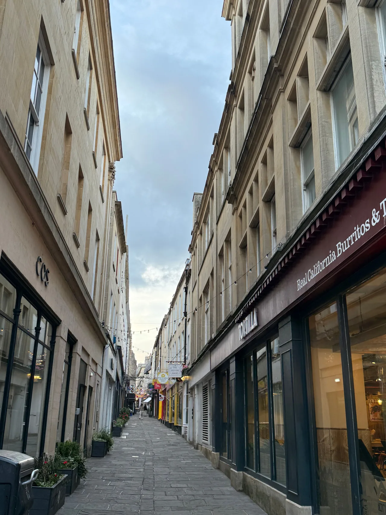 Uma rua pedestre estreita no centro de Bath ao anoitecer, ladeada por edifícios georgianos de pedra, pequenas lojas e fileiras de luzes.