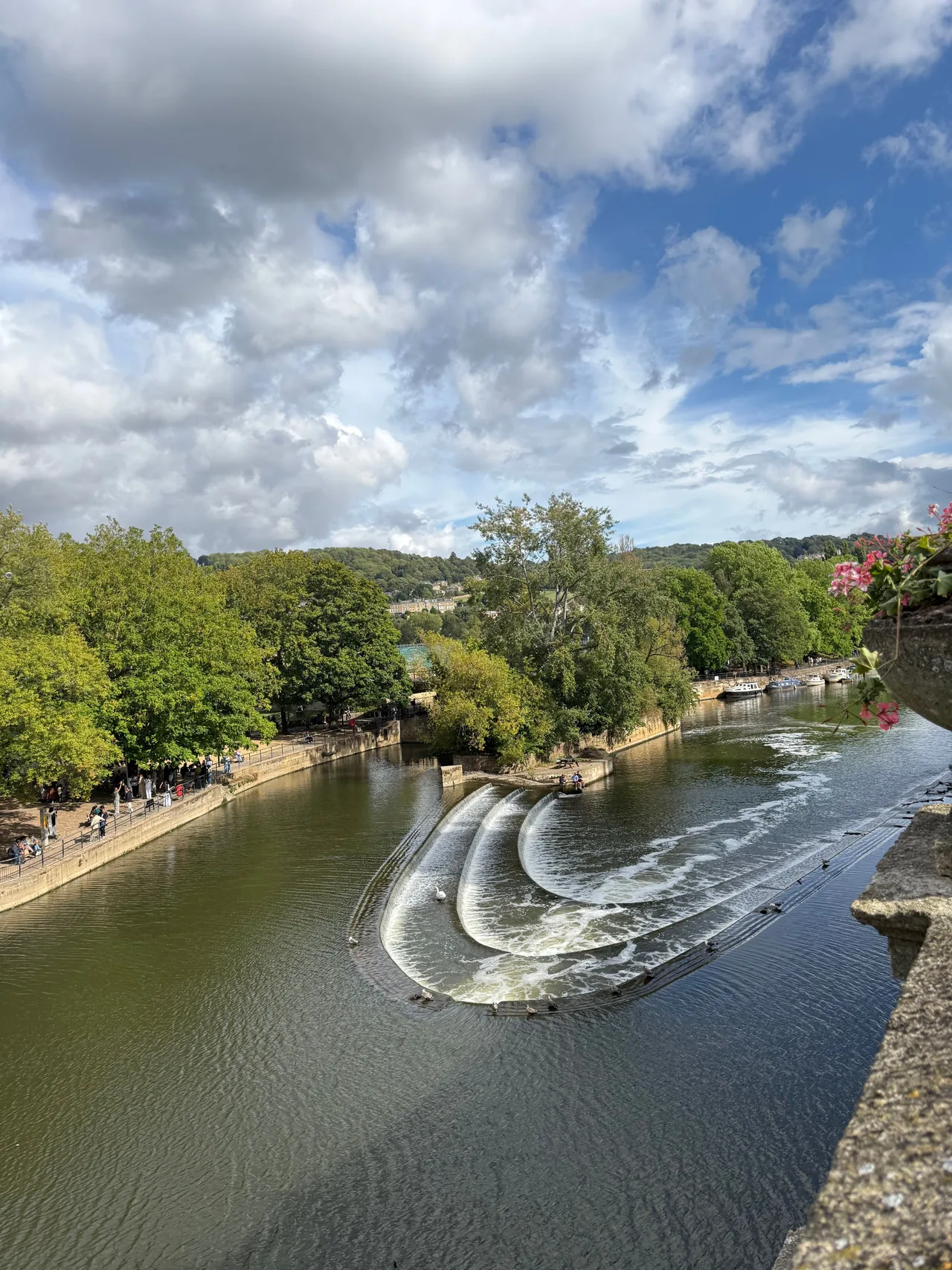 Pulteney Weir no Rio Avon em Bath, com seus degraus curvos de água branca, margens arborizadas e um caminho à beira do rio.