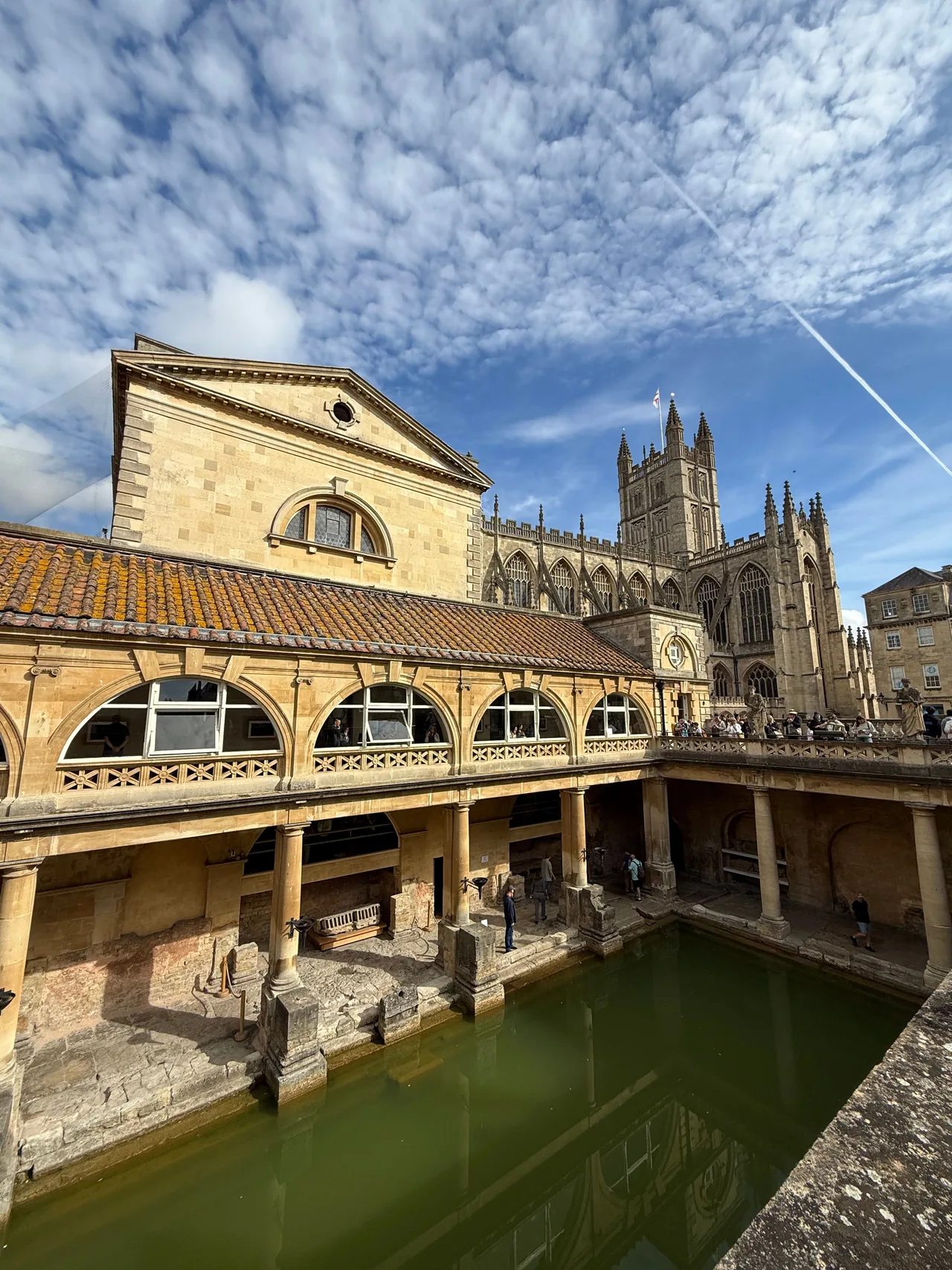 O Grande Banho nos Banhos Romanos em Bath, com água verde, colunas de pedra, o terraço revestido de estátuas e a Abadia de Bath ao fundo sob um céu azul.
