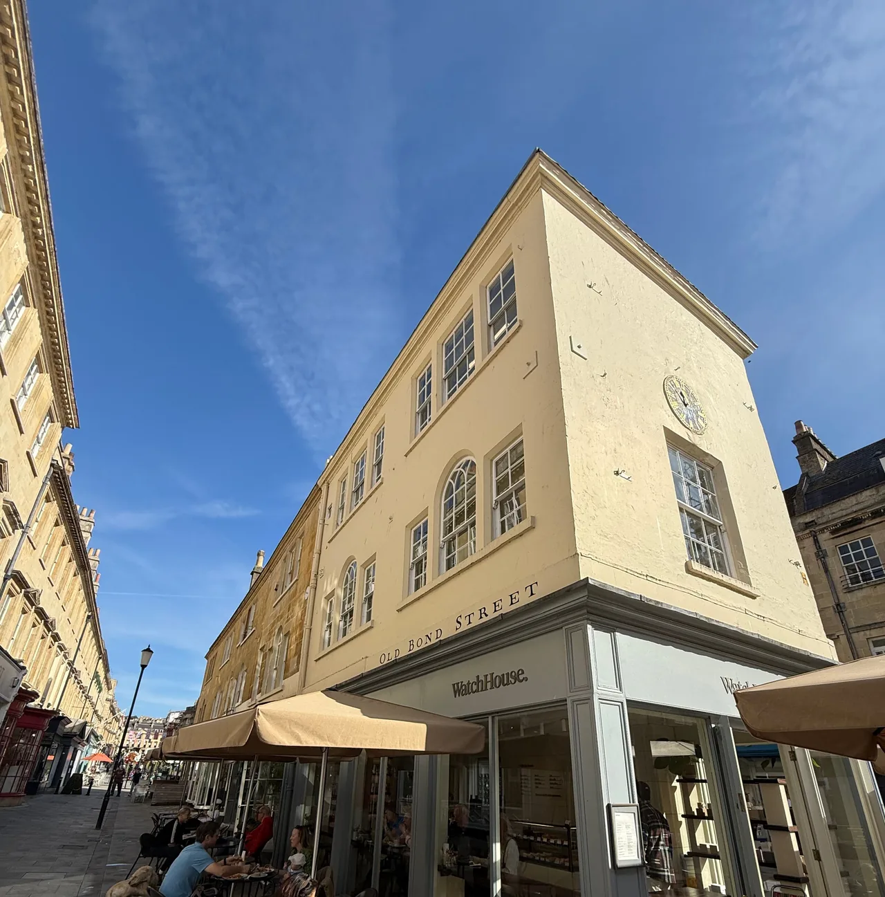 A cafeteria WatchHouse na esquina da Old Bond Street em Bath, um edifício georgiano creme com relógio, guarda-sóis e mesas ao ar livre sob um céu azul.