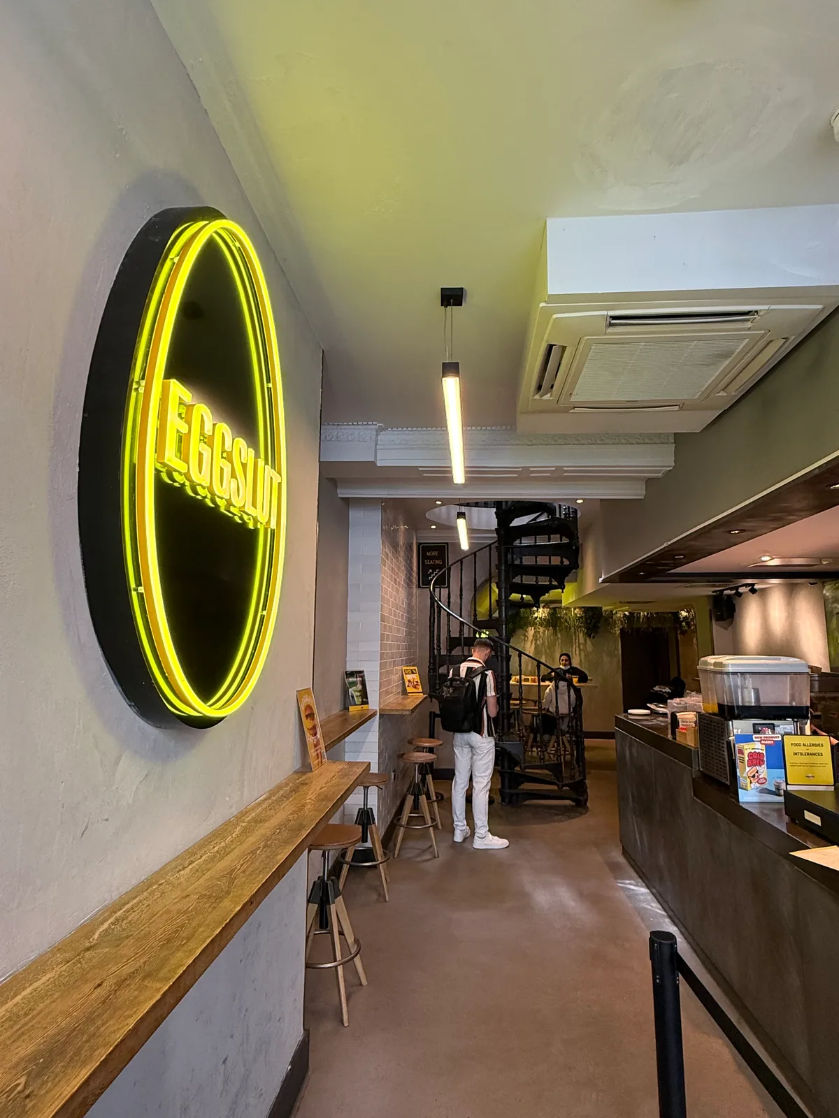Inside Eggslut Fitzrovia, a yellow neon eggslut oval sign above a counter bench with a spiral staircase up to the seating