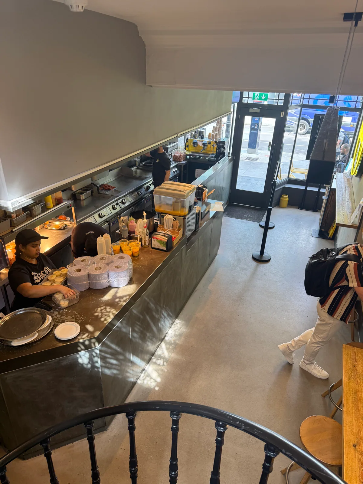 The open kitchen at Eggslut Fitzrovia seen from the stairs, staff cooking behind the steel counter with the street through the window