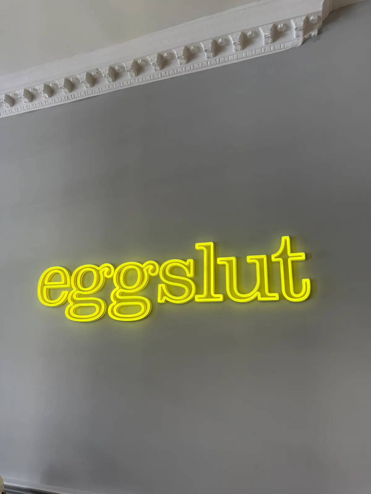 Yellow eggslut neon sign on a grey wall beneath ornate white period cornicing inside the Fitzrovia branch