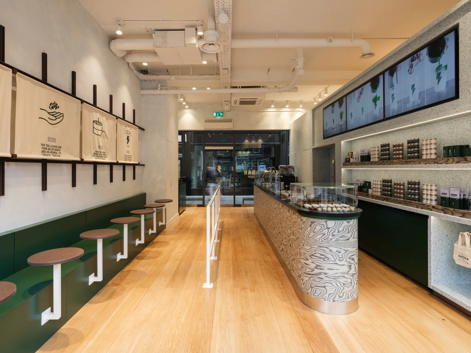 London matcha bar interior — light wood floor, green built-in seating with white stool stems, terrazzo counter with woodgrain inlay, ceremonial matcha tins lined up on the wall, screens showing the day's drinks