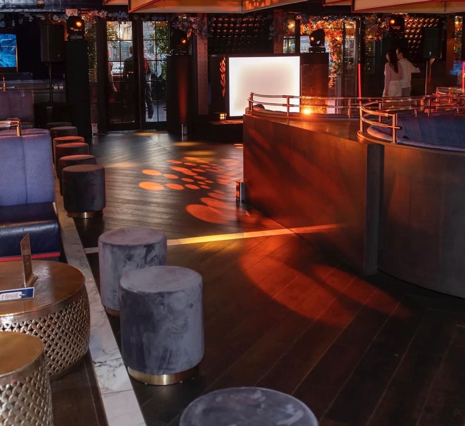 A moody Soho lounge interior at night with low lighting, gobo light patterns on the floor, navy velvet stools, and brass details