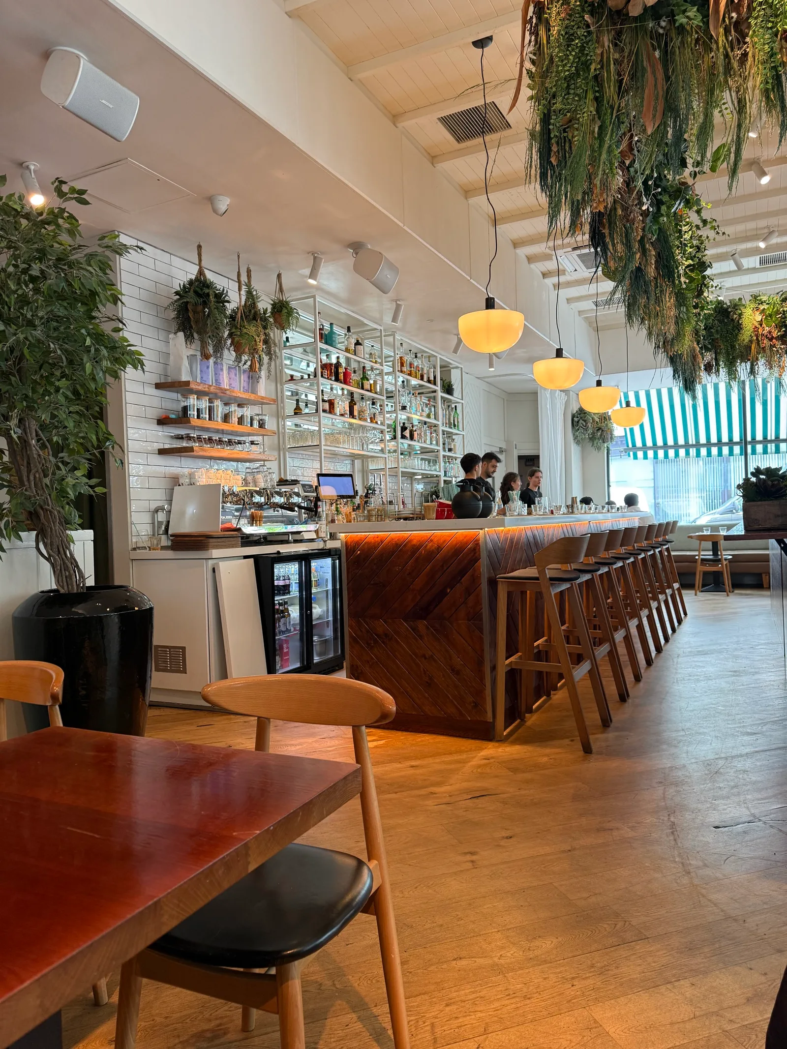 Sunday in Brooklyn interior on Westbourne Grove — long bar counter with wooden stools, hanging amber pendant lights, kitchen pass at the back, hanging plants and white tiled walls