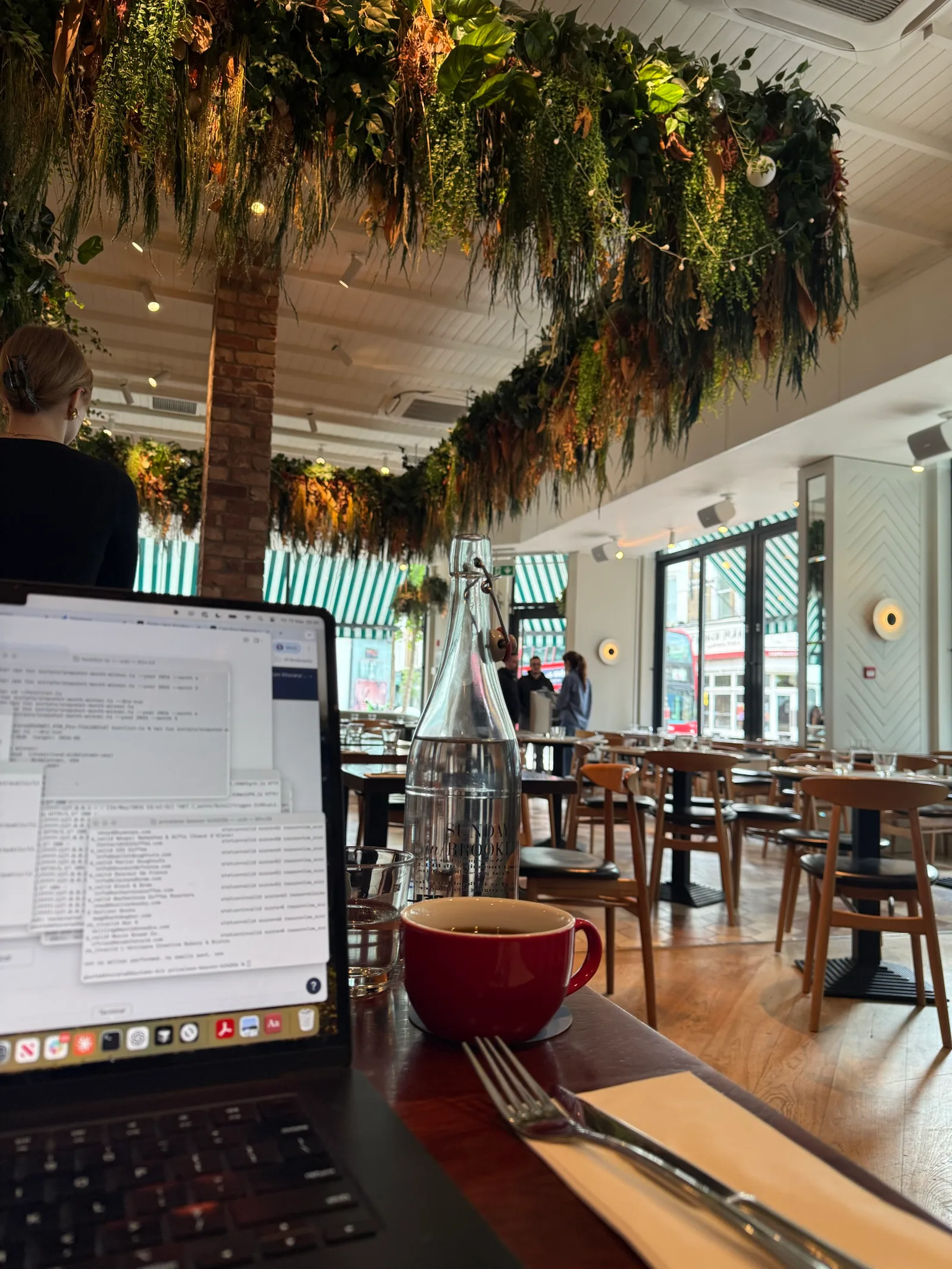 Working at Sunday in Brooklyn on Westbourne Grove — laptop open on a wooden bar top, red Le Creuset-style mug of drip coffee, tall water glass, hanging plants in soft light, the recognisable view I sign in to for three hours of focused work
