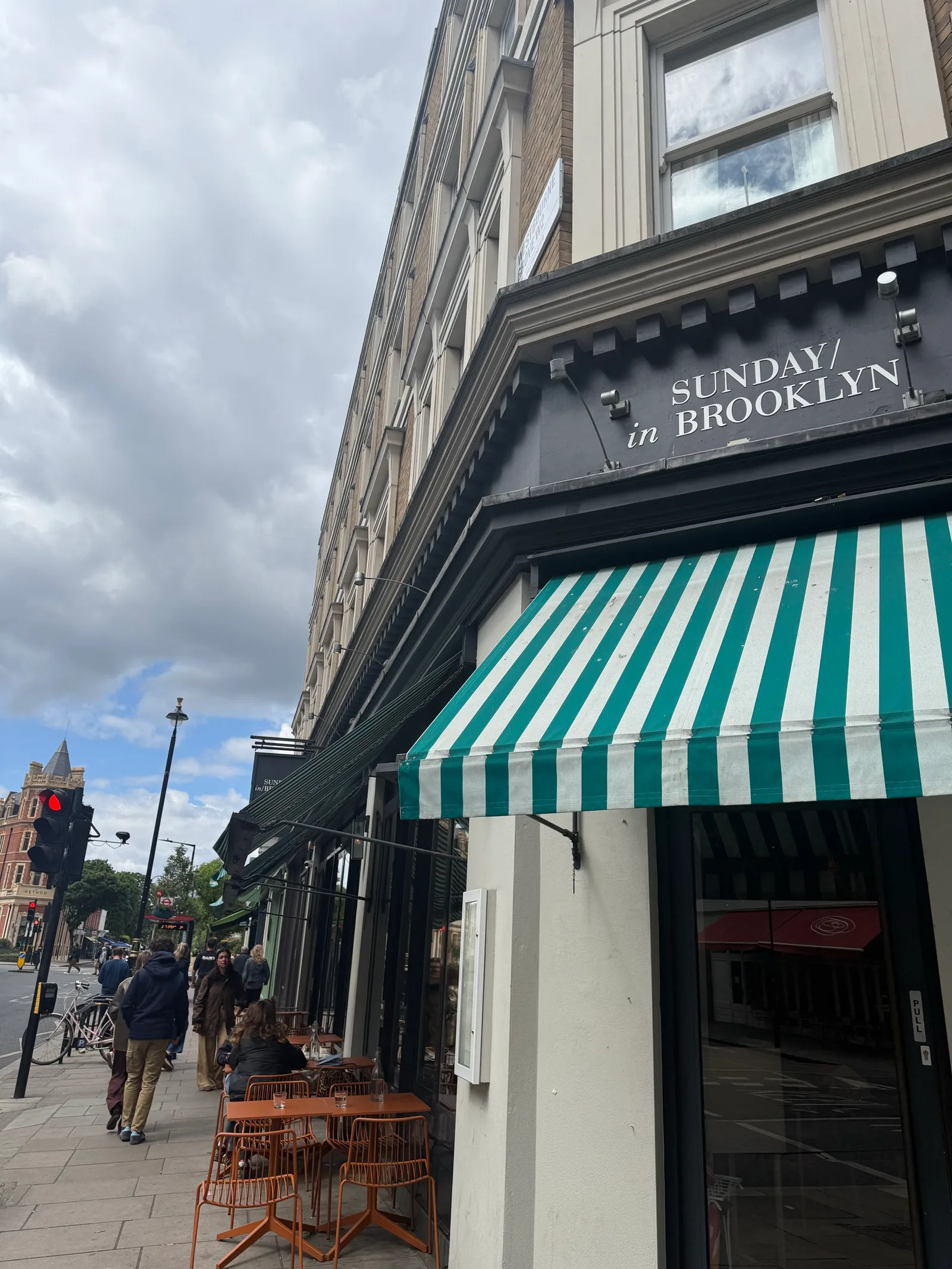 Sunday in Brooklyn storefront on Westbourne Grove, Notting Hill — green-and-white striped awning with the SUNDAY in BROOKLYN sign, sidewalk seating with orange wire chairs, classic Notting Hill stucco building above